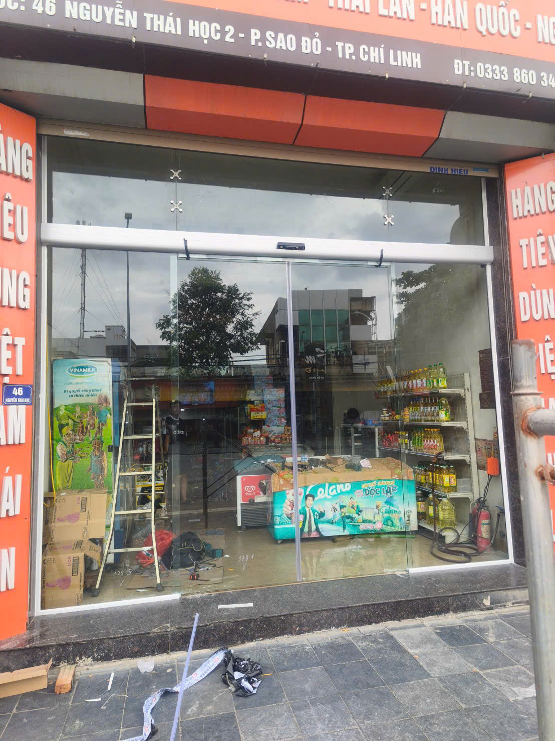 Cửa hàng tạp hóa với cửa kính trưng bày kem que Algida và kệ dầu ăn, phía trước đang thi công lắp đặt